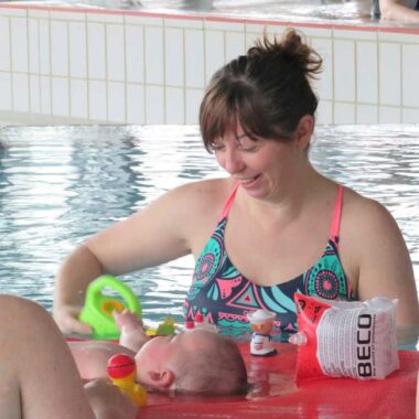 Eine lächelnde Frau in einem bunten Badeanzug beugt sich über ein Baby im Wasser und interagiert mit Schwimmspielzeug, das auf einer roten Unterlage liegt.