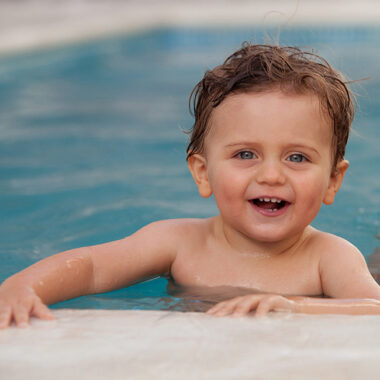 Ein glücklich lachender kleiner Junge mit nassen, braunen Haaren und strahlend blauen Augen lehnt sich am Beckenrand eines Schwimmbades ab.