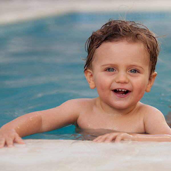 Ein glücklich lachender kleiner Junge mit nassen, braunen Haaren und strahlend blauen Augen lehnt sich am Beckenrand eines Schwimmbades ab.